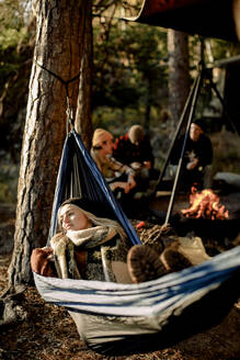 Junge Frau entspannt sich in einer Hängematte mit Freunden beim Zelten im Hintergrund - MASF34211