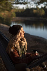 Young woman with eyes closed holding coffee cup while sitting on hammock - MASF34212