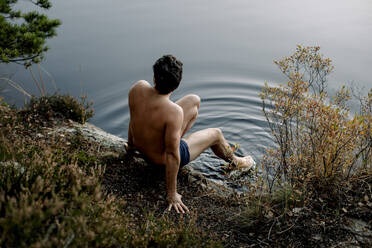 Shirtless man sitting near lake with legs dipped in water - MASF34228