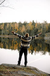 Rear view of carefree man with arms outstretched standing on rock against lake - MASF34238