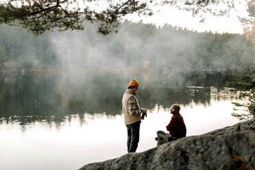 Male friends talking to each other on rock near lake during staycation - MASF34246