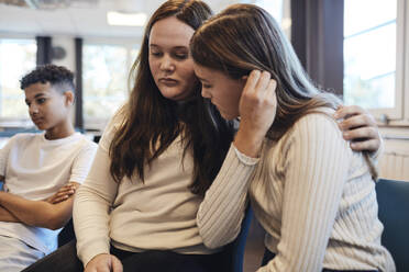 Sad teenage girl sitting with arm around female friend while consoling her in group therapy - MASF34268