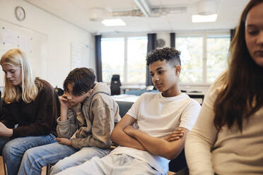 Nachdenklicher Teenager sitzt mit verschränkten Armen inmitten von Freunden im Klassenzimmer während einer Gruppentherapie - MASF34269