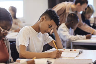 Bored male student writing in book while sitting by female friend at desk - MASF34280