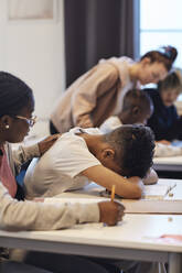 Teenager-Mädchen, das einen deprimierten männlichen Schüler tröstet, der seinen Kopf im Klassenzimmer auf den Schreibtisch legt - MASF34282