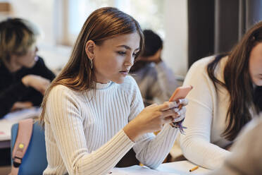 Teenager-Mädchen mit braunen Haaren, das ein Smartphone benutzt und neben einer Freundin am Schreibtisch im Klassenzimmer sitzt - MASF34286