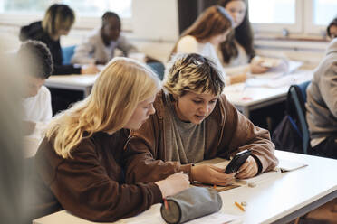 Teenager-Mädchen teilt sich ein Smartphone mit einer blonden Freundin, die am Schreibtisch im Klassenzimmer sitzt - MASF34289