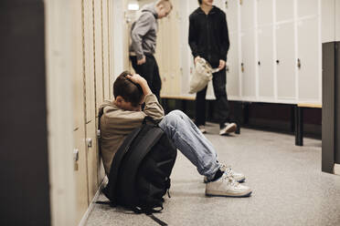Depressiver Junge in voller Länge mit Rucksack im Schulkorridor sitzend - MASF34303