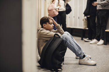 Seitenansicht eines traurigen Teenagers, der mit einem Rucksack im Schulkorridor sitzt - MASF34304
