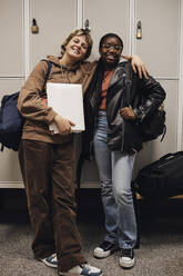 Full length portrait of smiling teenage girl standing with arm around female friend against locker in school - MASF34309