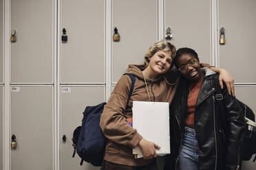 Portrait of smiling female friends standing together against locker in school - MASF34310