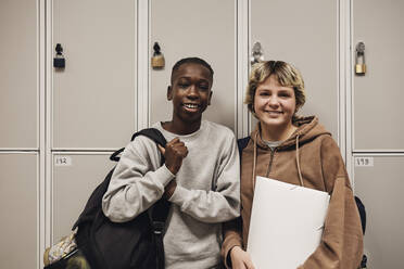 Portrait of smiling male and female teenage friends standing against locker in school - MASF34312