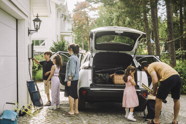 Familie beim Aufladen des Gepäcks im Kofferraum eines Elektroautos vor dem Haus - MASF34360