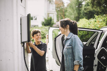 Smiling boy holding cable while looking at mother standing near electric car - MASF34363