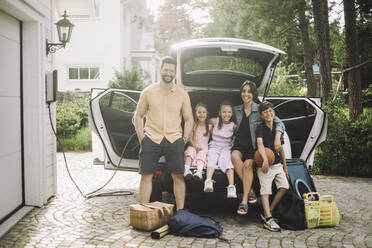 Porträt einer glücklichen Familie, die im Kofferraum eines Elektroautos vor dem Haus sitzt - MASF34370