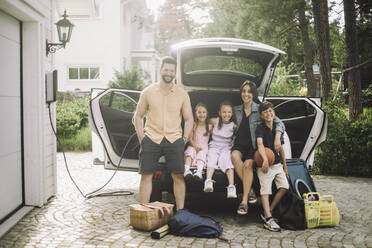 Portrait of happy family sitting in electric car trunk outside house - MASF34370