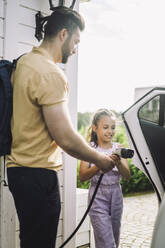 Seitenansicht eines Vaters, der seiner Tochter beibringt, wie man ein Elektroauto auflädt - MASF34381