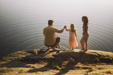 Vater hält die Hand seiner Tochter, während er an einem sonnigen Tag in der Nähe eines Sees hockt - MASF34410