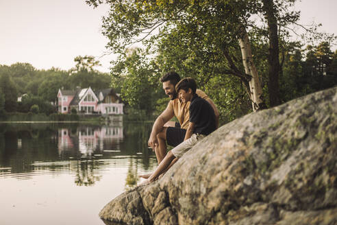 Vater und Sohn sitzen auf einem Felsen am See - MASF34412