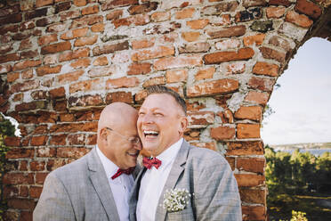 Fröhliches frisch verheiratetes homosexuelles Paar, das sich an einer Backsteinmauer vergnügt - MASF34421