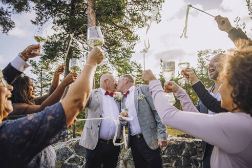 Neuvermähltes homosexuelles Paar, das sich küsst, während Freunde und Familie ihnen bei der Hochzeit zujubeln - MASF34427