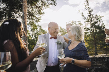 Lächelnde ältere Frau und Bräutigam mit Weingläsern, die sich bei der Hochzeit umarmen - MASF34429