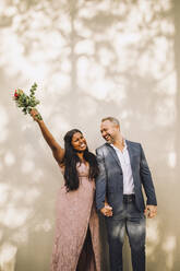 Glückliche Braut mit Blumenstrauß und erhobener Hand neben dem Bräutigam an der Wand stehend - MASF34435
