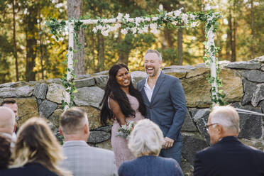 Glückliches neuvermähltes gemischtrassiges Paar, das die Gäste während der Hochzeitszeremonie ansieht - MASF34443