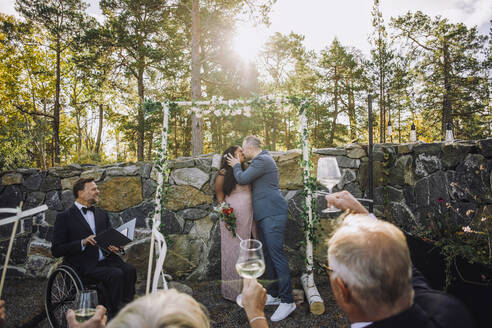 Neuvermähltes Paar küsst sich vor den Gästen, die bei der Hochzeitszeremonie Trinksprüche ausbringen, und einem Pfarrer mit Behinderung - MASF34445