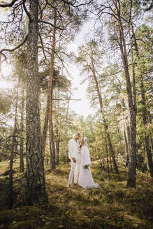 Mid Erwachsenen Braut hält Bouquet Blick auf Bräutigam stehend inmitten von Bäumen im Wald - MASF34449