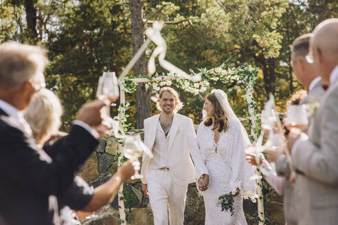 Neuvermähltes Paar im mittleren Erwachsenenalter, das sich an den Händen hält, während einer Hochzeitszeremonie inmitten der Gäste - MASF34459