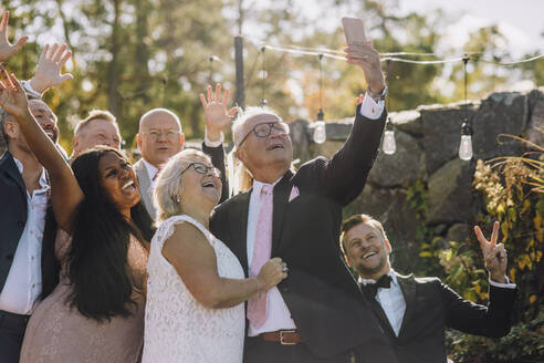 Glücklich frisch verheiratetes älteres Paar, das ein Selfie mit Familie und Freunden über ein Smartphone macht - MASF34468