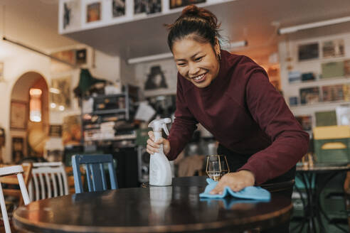 Glückliche Kaffeehausbesitzerin mit Sprühflasche und Lappen beim Reinigen des Tisches im Café - MASF34477