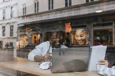 Happy female coffee shop owners discussing over laptop seen through window - MASF34490
