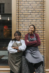 Portrait of smiling female cafe owners wearing aprons standing with arms crossed outside coffee shop - MASF34491