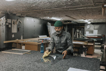 Älterer Unternehmer mit Strickmütze, der an einer Werkbank Glas mit einem Lappen reinigt - MASF34505