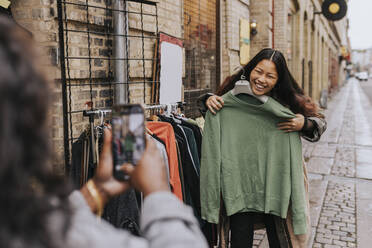 Mature woman photographing cheerful female friend trying on clothes at street - MASF34541