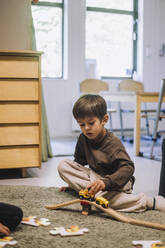 Junge spielt mit Spielzeugeisenbahn, während er auf dem Teppich im Kindergarten sitzt - MASF34549