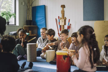 Happy multiracial male and female students enjoying while playing drum kit in child care center - MASF34554