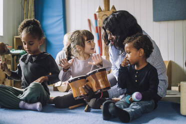 Female teacher teaching girl to play drum kit by classmates in day care center - MASF34556