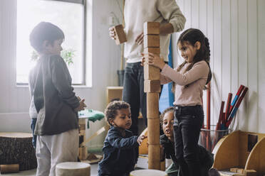 Weibliche und männliche Schüler stapeln Holzspielzeugklötze mit einem männlichen Lehrer in einer Kindertagesstätte - MASF34561