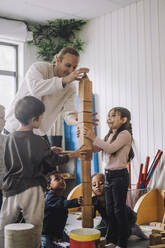 Male teacher guiding children stacking wooden toy blocks at kindergarten - MASF34562