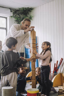 Männlicher Lehrer, der Kinder beim Stapeln von Holzklötzen im Kindergarten anleitet - MASF34562