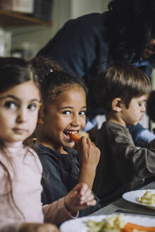 Glückliches Mädchen isst Karotte zum Frühstück mit Klassenkameraden im Kindergarten - MASF34571