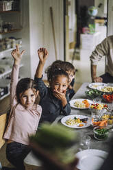 Glückliche Mädchen und Jungen mit erhobenen Händen am Esstisch beim Frühstück im Kindergarten - MASF34573