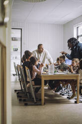 Female and male teachers talking with children having breakfast at dining table - MASF34577