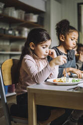 Girl having pasta for breakfast with female classmate in child care center - MASF34580