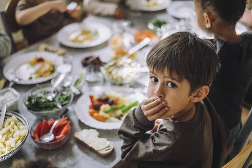 Porträt eines Jungen, der den Mund mit der Hand bedeckt, während das Frühstück auf dem Tisch einer Kindertagesstätte steht - MASF34582