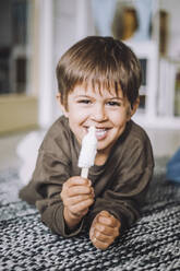 Porträt eines lächelnden Jungen, der auf einem Teppich liegend ein Eis isst - MASF34597
