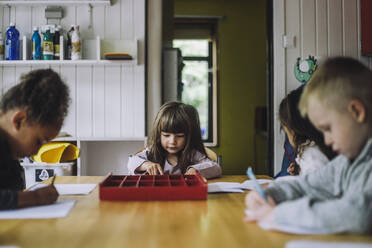 Männliche und weibliche Schüler verschiedener Rassen schreiben an einem Tisch im Kindergarten auf Papier - MASF34616